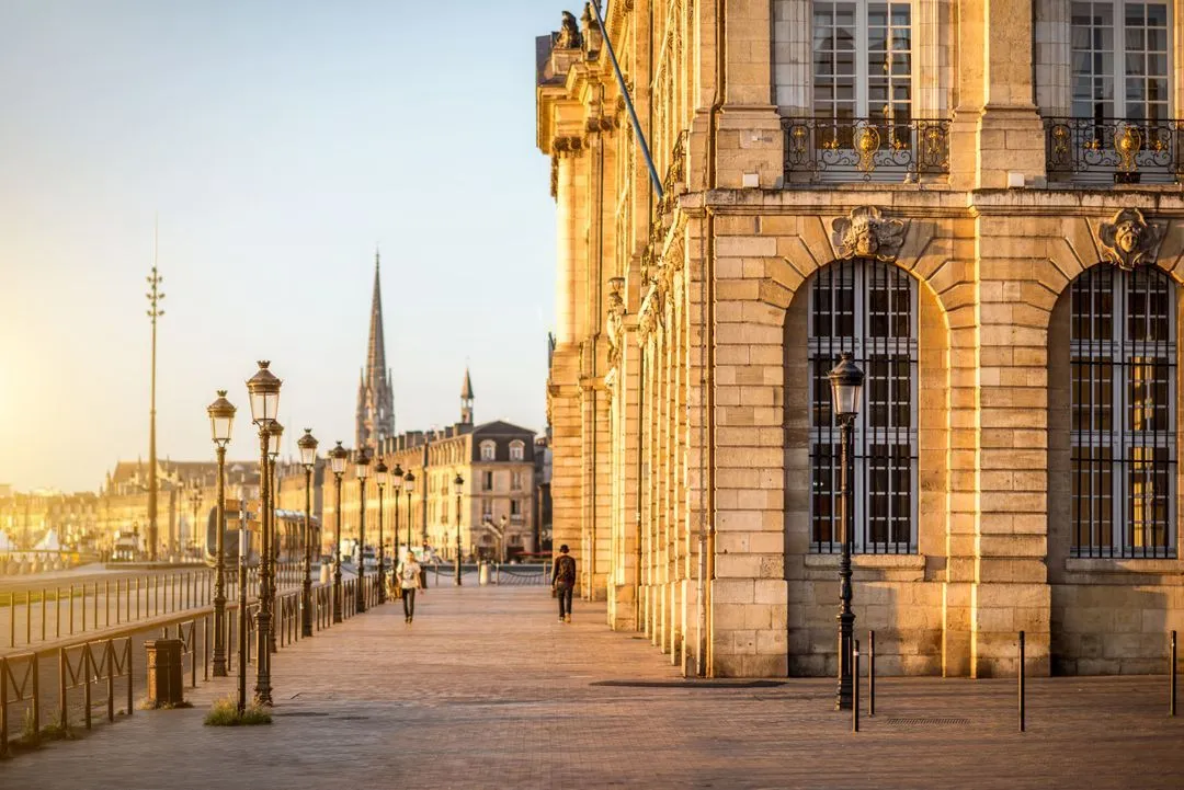 Vue ensoleillée des quais de Bordeaux avec des lampadaires anciens, des passants et la façade d’un bâtiment historique en pierre, sur fond de ciel dégagé et de la flèche de l’église Saint-Michel à l’horizon.